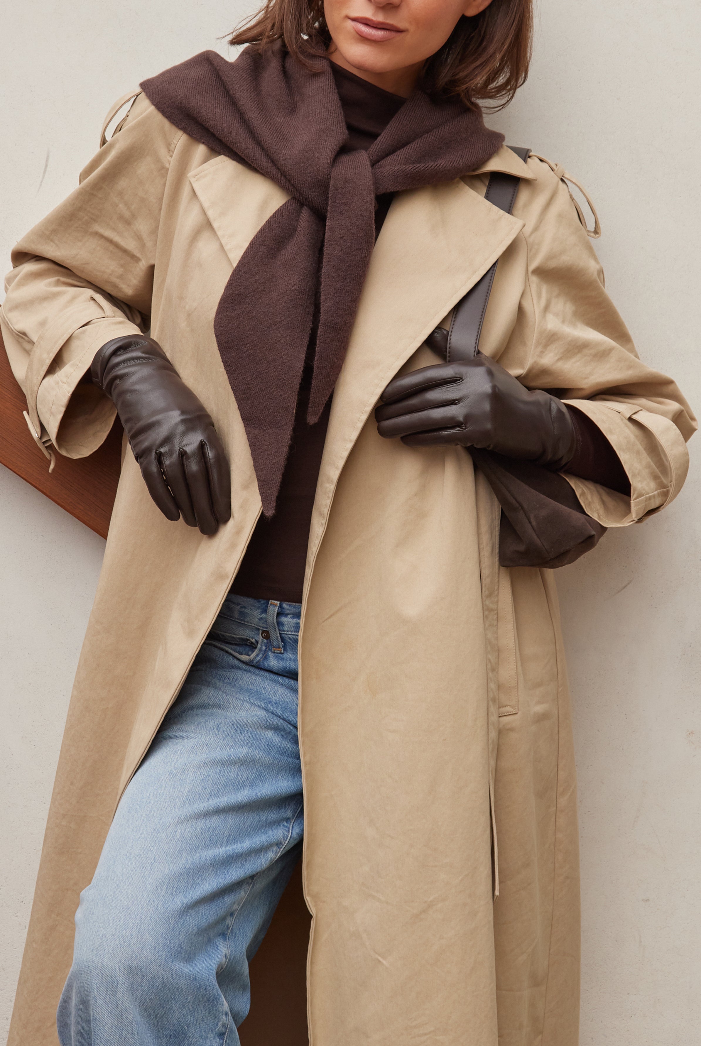 Person wearing a beige trench coat, brown scarf, brown gloves, and blue jeans against a white background