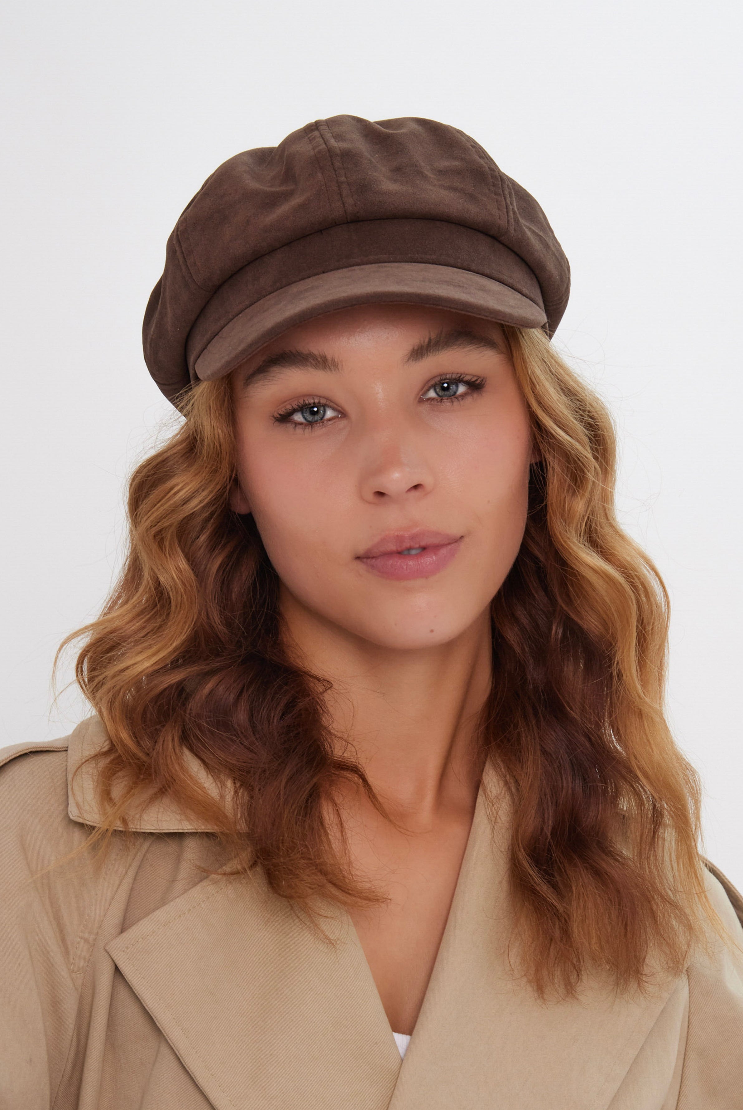 Woman wearing a brown suede baker boy cap and beige coat on a white background