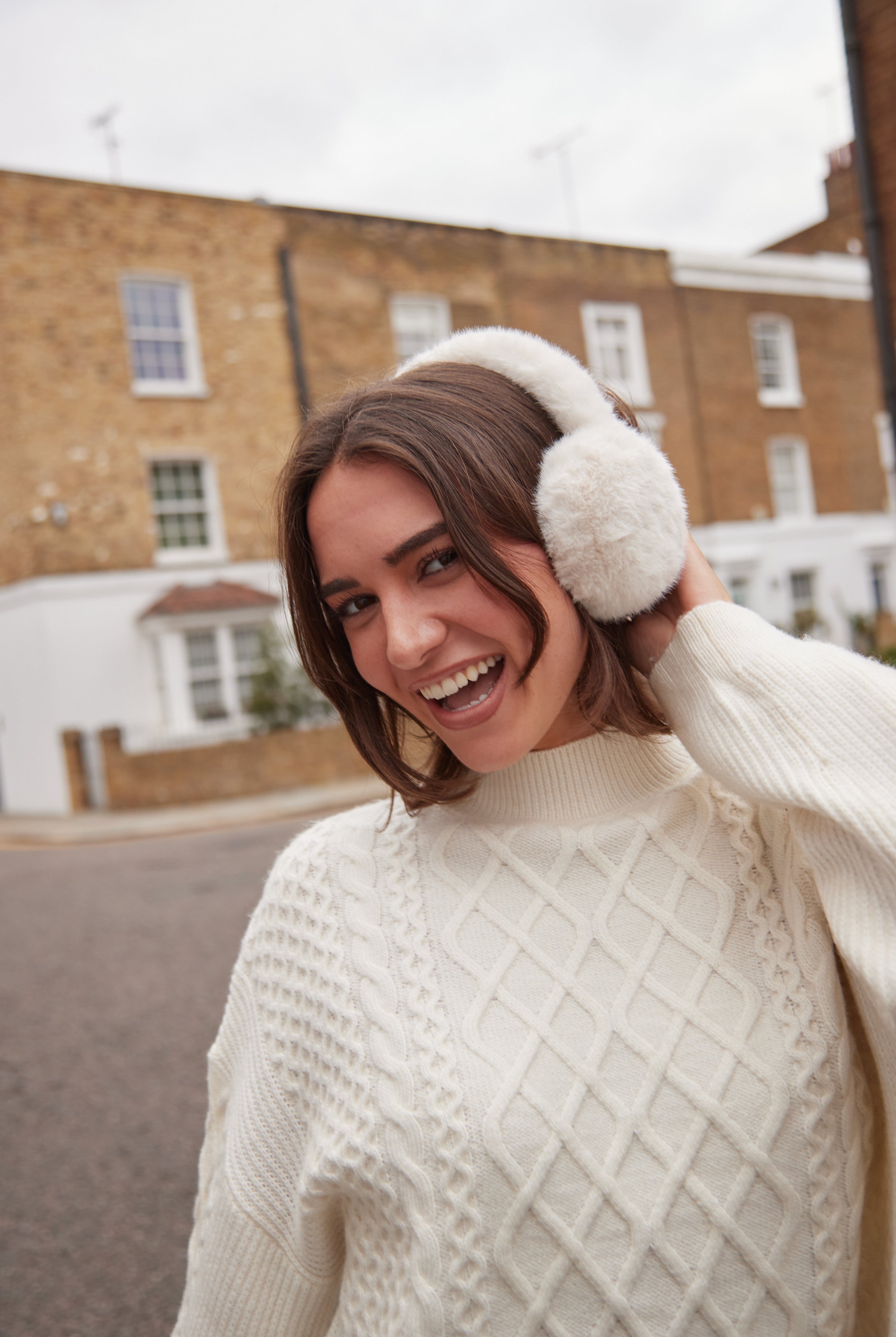 Woman wearing white ear muffs and a cream sweater outdoors.