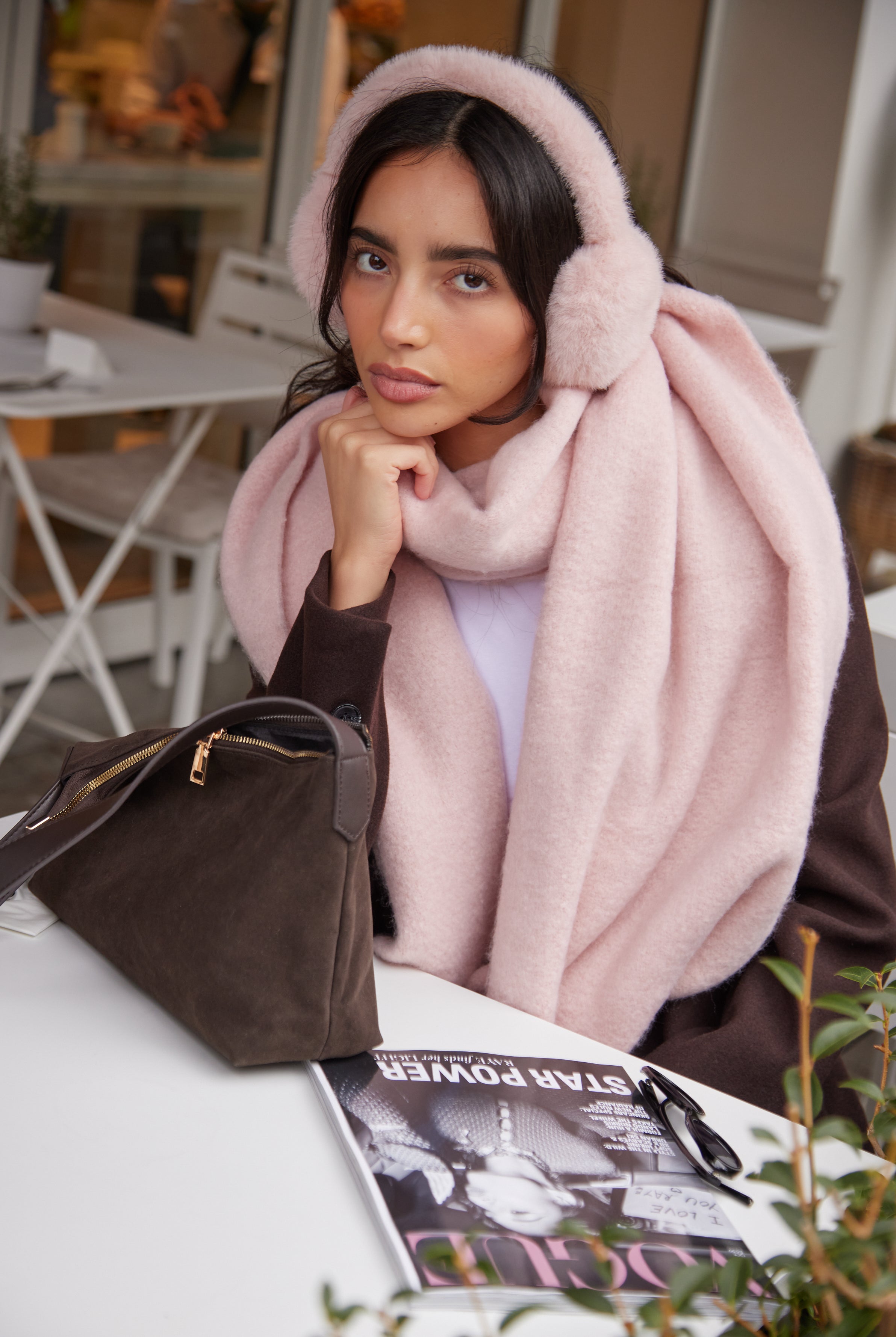 Woman wearing a pink scarf pink ear muffs sitting at a table with a handbag.