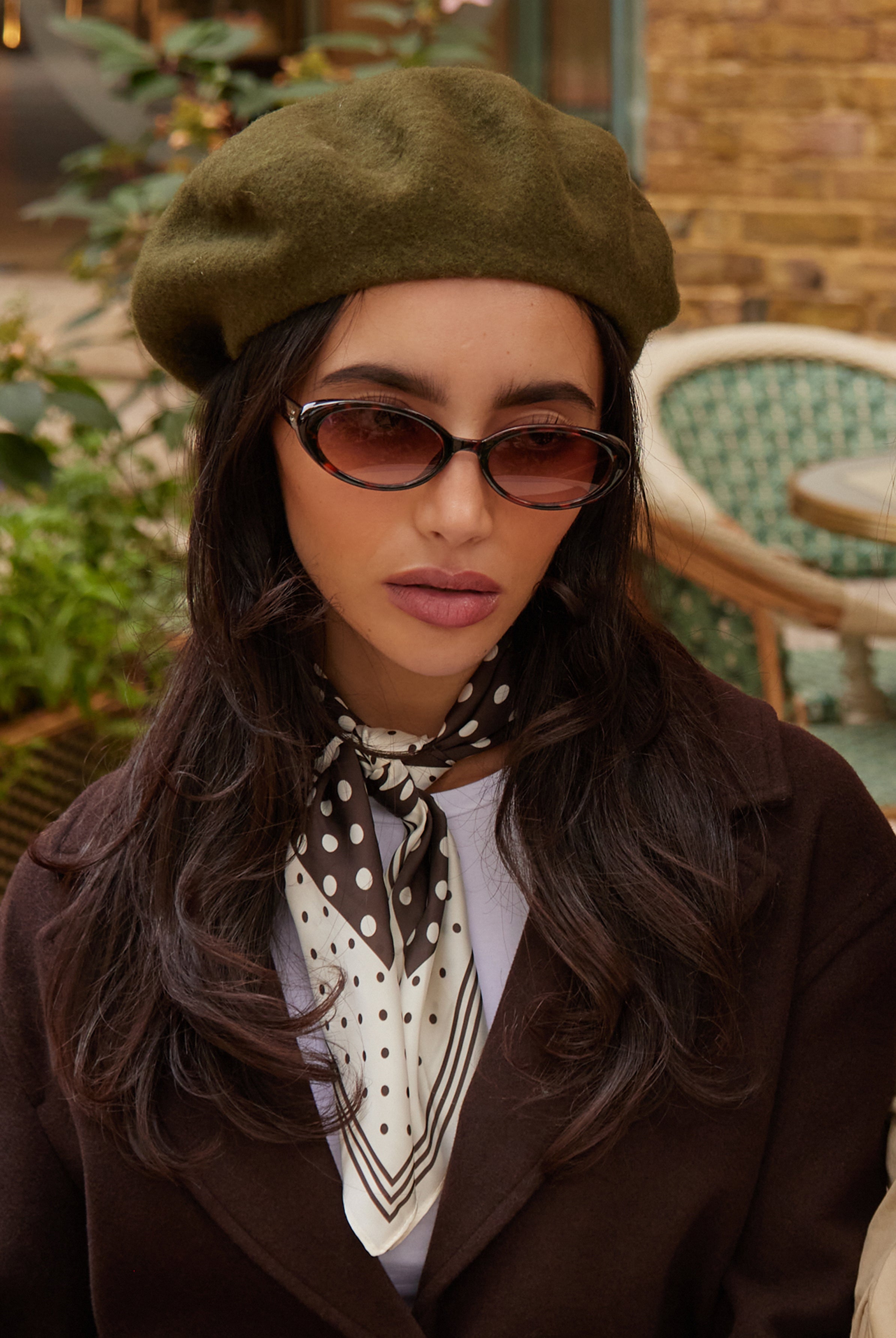 Woman wearing a green beret, sunglasses, and a brown coat with a polka dot scarf in an outdoor setting.