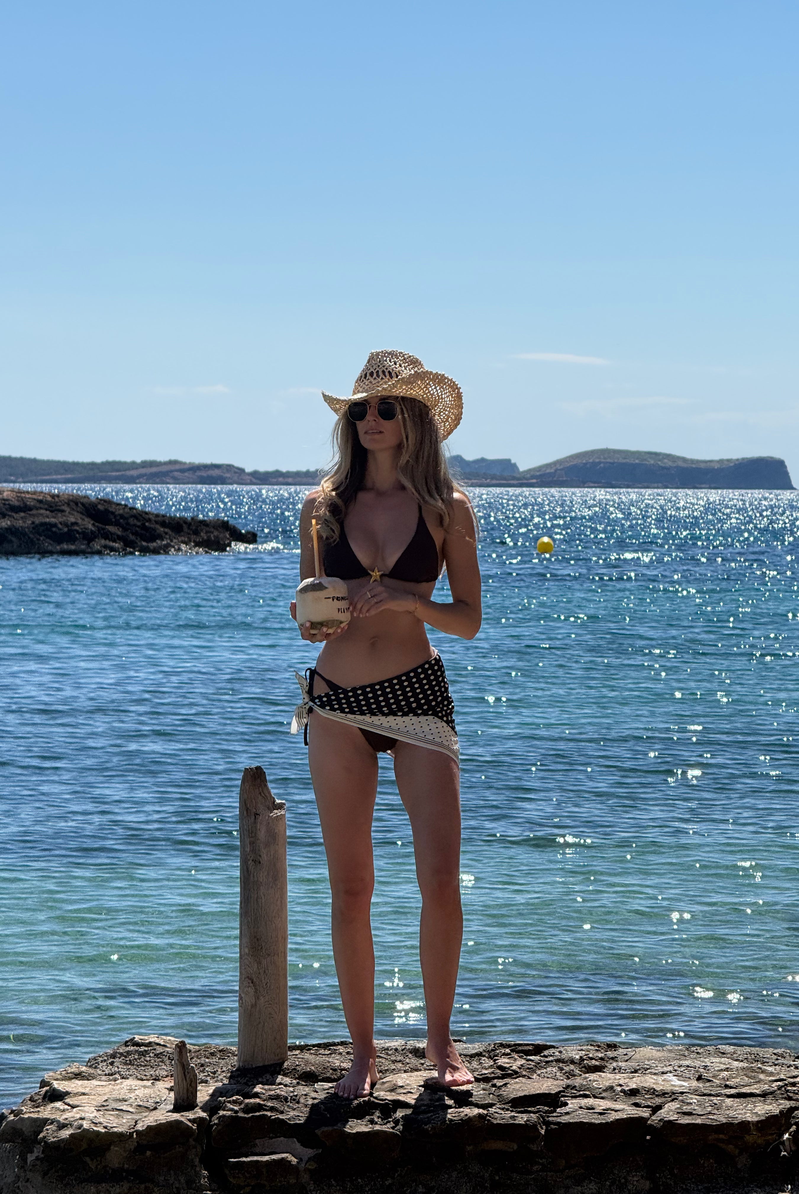 Woman in a bikini and hat and polka dot satin scarf standing on rocks by the ocean