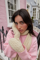 Woman wearing a pink top and cream gloves and scarf in front of a pink building.