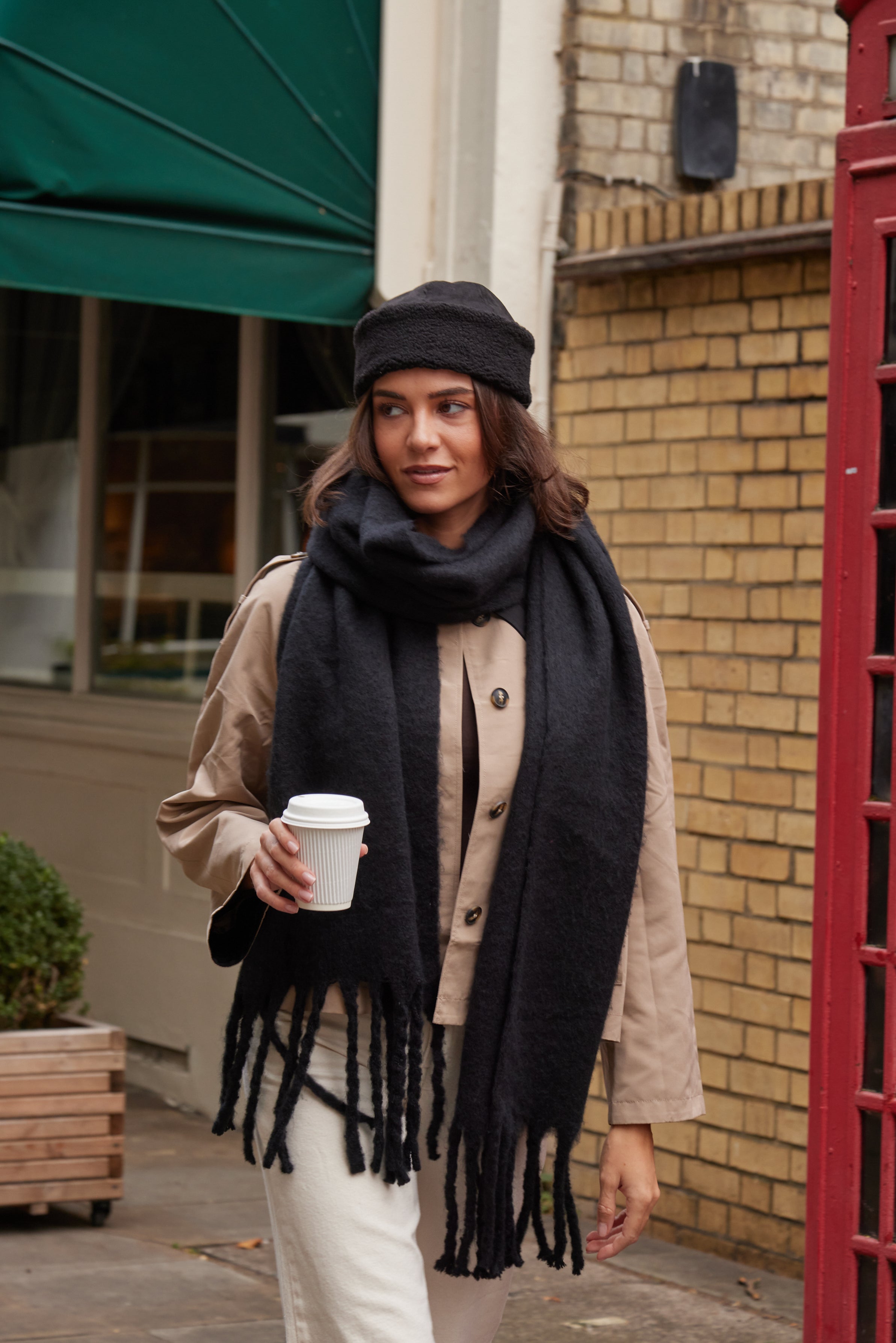 Woman wearing a black scarf and hat, holding a coffee cup, standing outside a building.