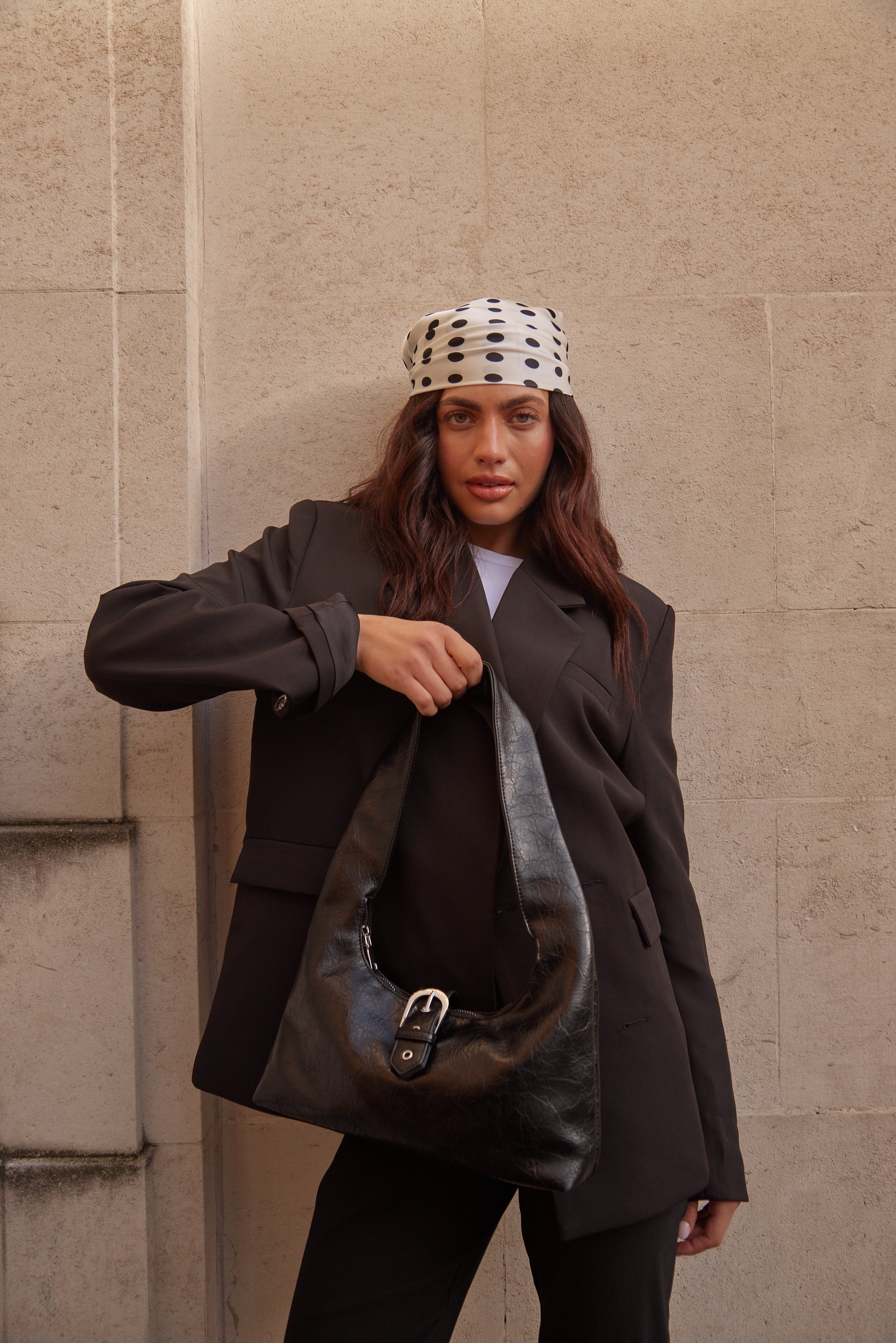 Person wearing a black coat with a black shoulder bag and headscarf against a beige wall
