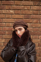 Person wearing a brown leather coat with fur mittens and hat against a brick wall.