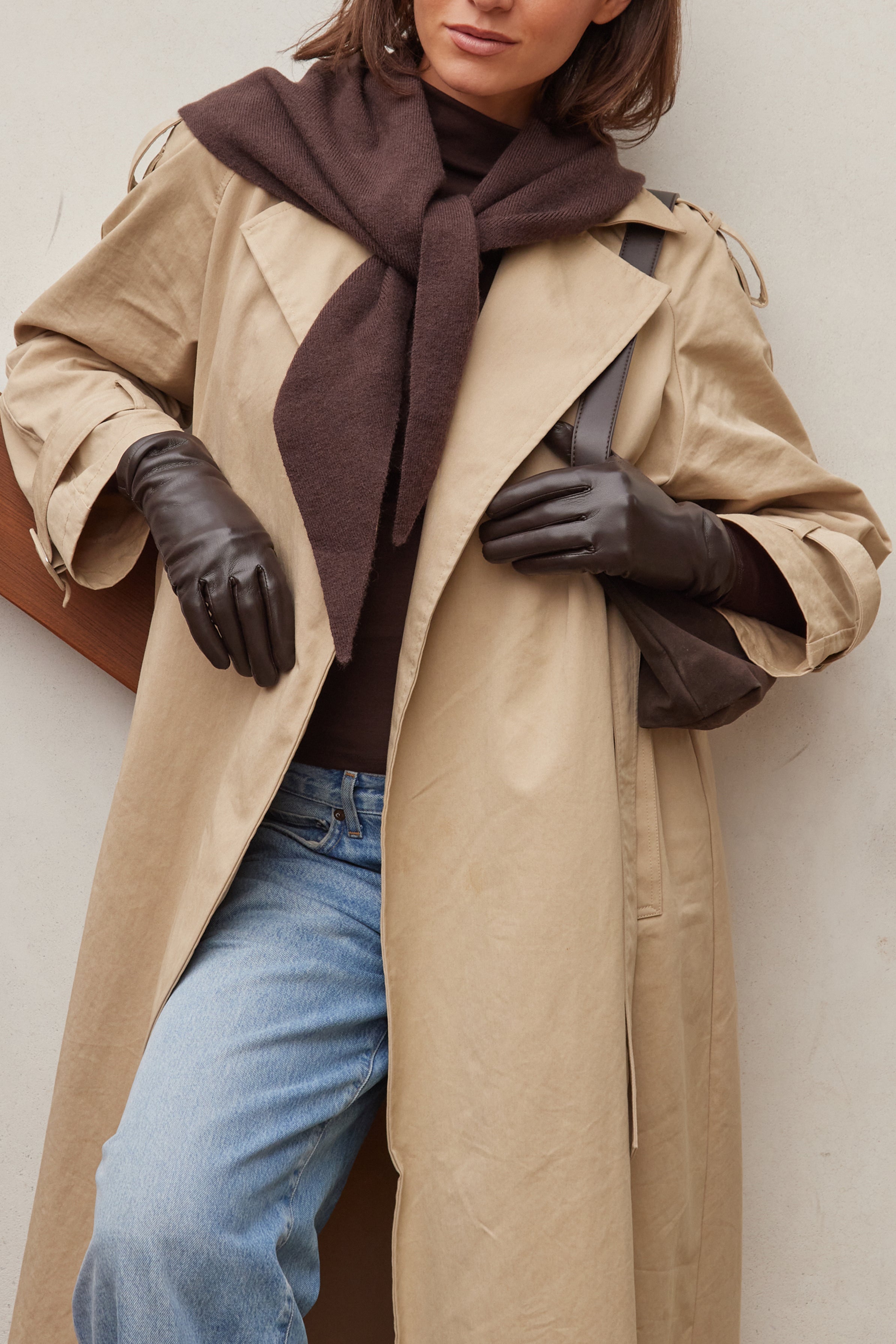 Person wearing a beige trench coat, brown scarf, brown gloves, and blue jeans against a white background