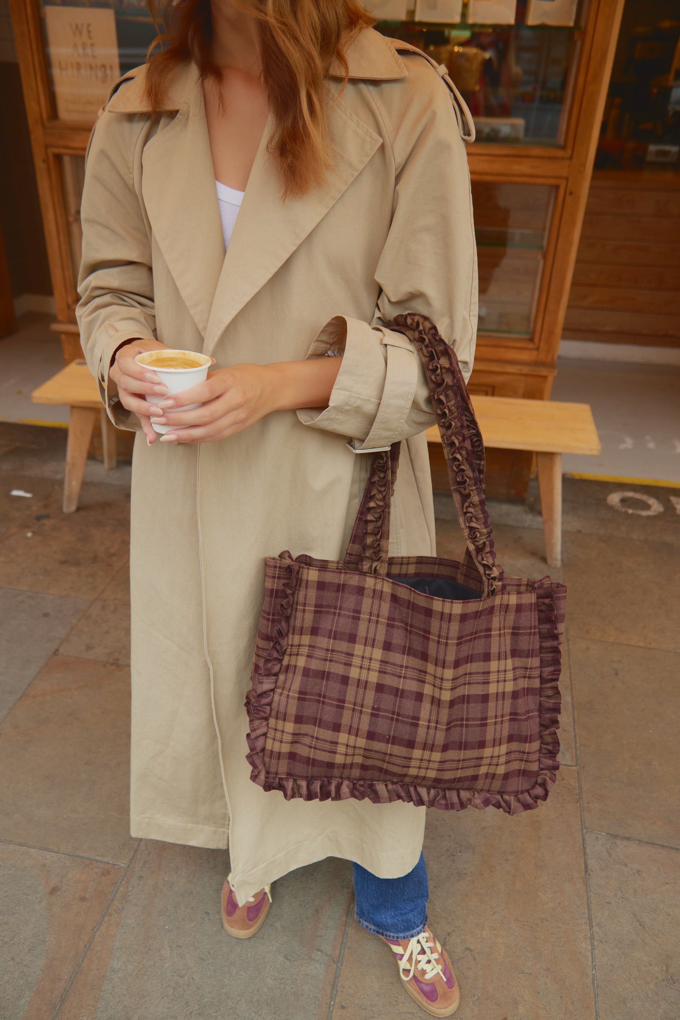 Person wearing a beige trench coat holding a plaid bag and a cup, standing indoors.