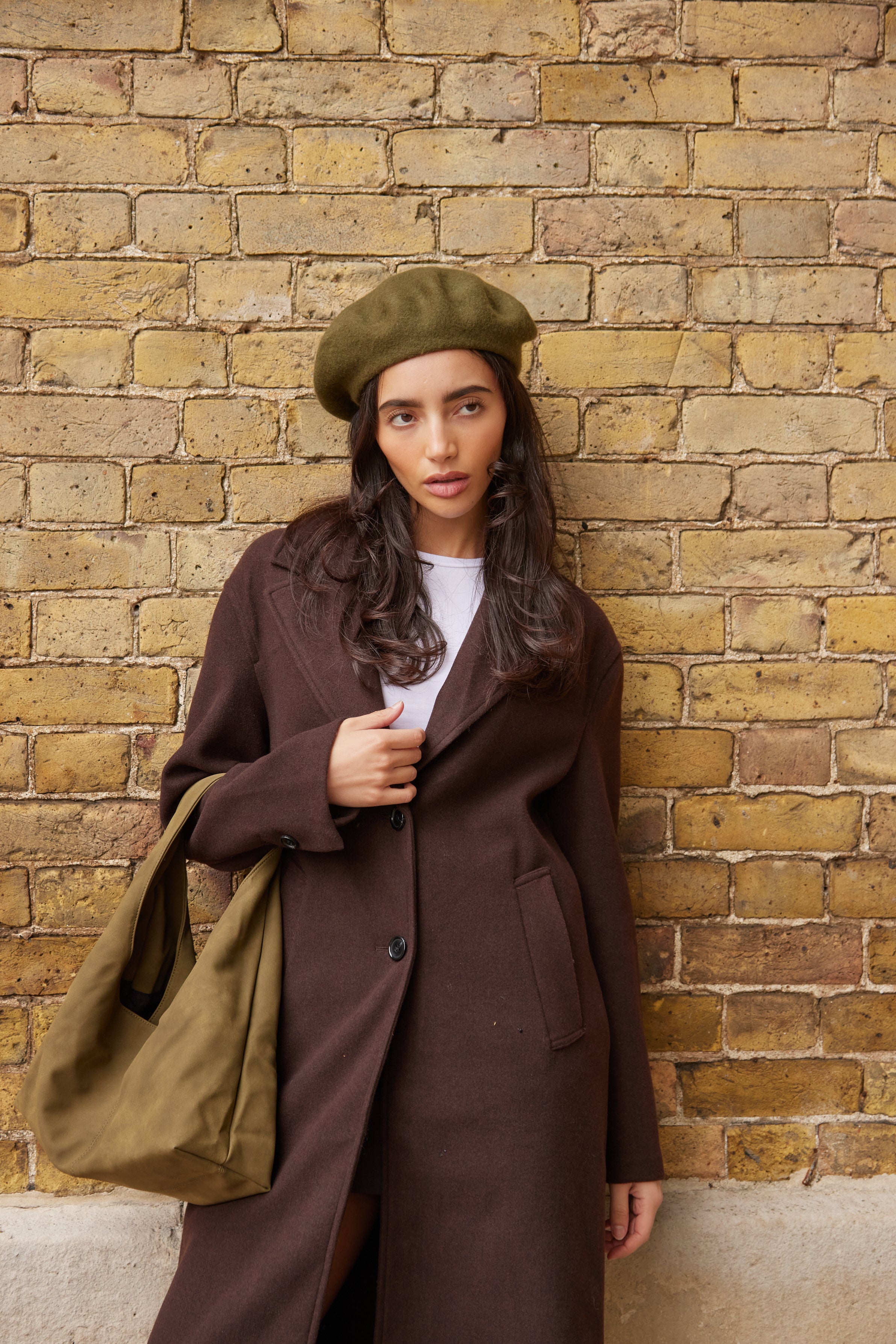 Woman in a brown coat and green beret and green bag standing against a brick wall