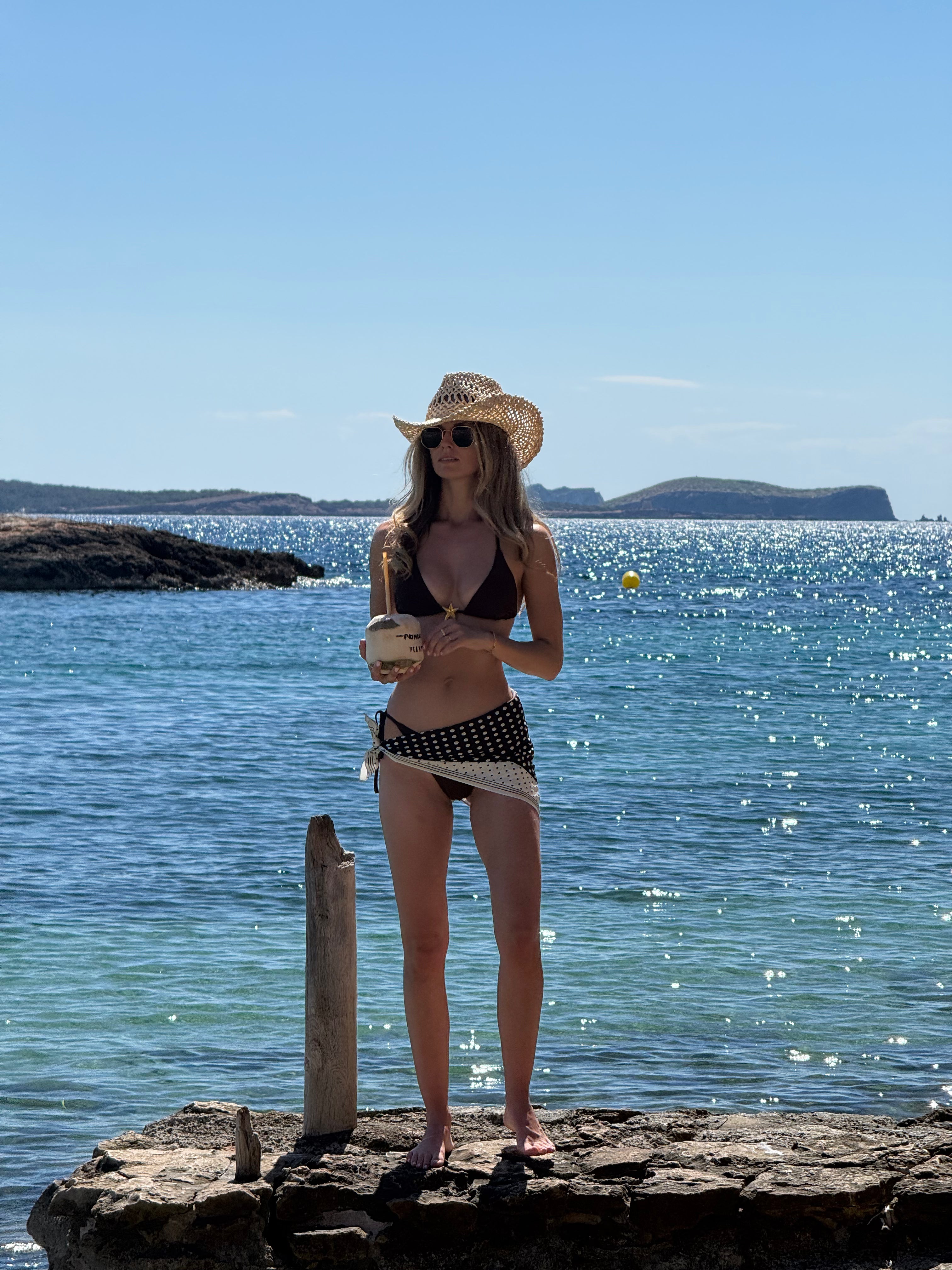 Woman in a bikini and hat and polka dot satin scarf standing on rocks by the ocean
