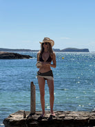 Woman in a bikini and hat and polka dot satin scarf standing on rocks by the ocean
